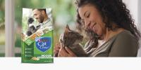 A man smiles while snuggling a grey tabby cat in a sunny room; a bag of Purina Cat Chow Naturals Indoor sits to the left