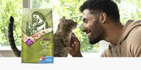 A man smiles as he pets a grey tabby cat in a sunny room; a bag of Purina Cat Chow Naturals Indoor sits to the left