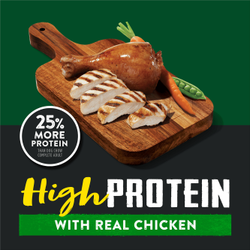 Slices of roasted chicken, carrots, and a pea pod sit on a wooden cutting board; the background is dark green and black. A circle badge left to the cutting board reads "25% more protein than Dog Chow Complete Adult." Text below the cutting board reads "high protein with real chicken"