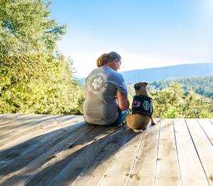 Person and service dog pug sitting on a wooden deck with a forest and hills in the background.