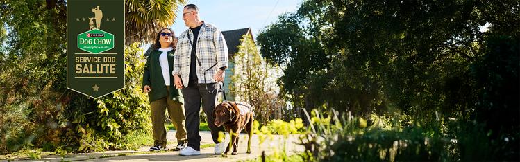 Two people walking a brown service dog in a leafy area with a banner stating "Purina Dog Chow Service Dog Salute."