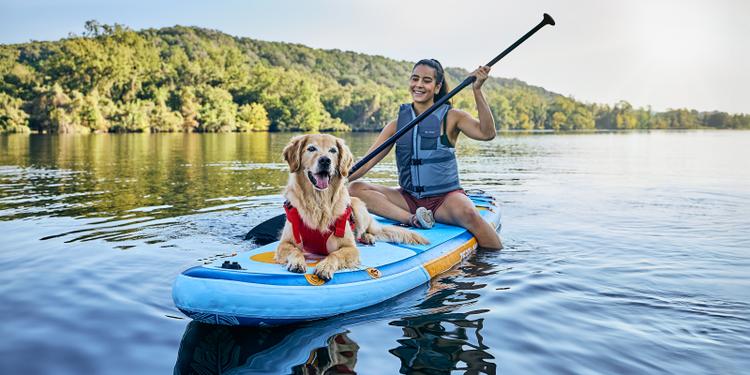 A young woman sits with her dog on a paddleboard in a lake.