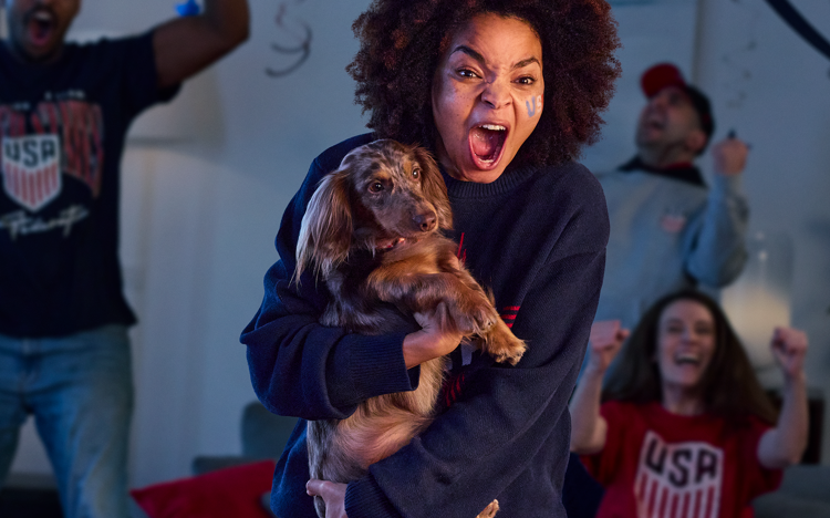 A woman holding a dachshund surrounded by a group of people cheering for US Soccer.