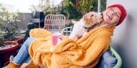 Una persona sentada en una silla de patio envuelta en una manta de lana acogedora mientras nieva. Su perro lame su cara.