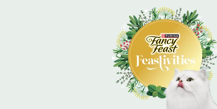 A circular gold Feastivities sign with green leaves and flowers and the white long haired Fancy Feast cat in the foreground