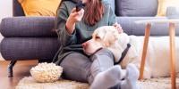 Woman sitting on floor in front of couch next to a bowl of popcorn with a white dog's head in her lap