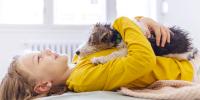 Una niña que viste suéter amarillo se acuesta de espaldas y abraza a un cachorro de aspecto triste