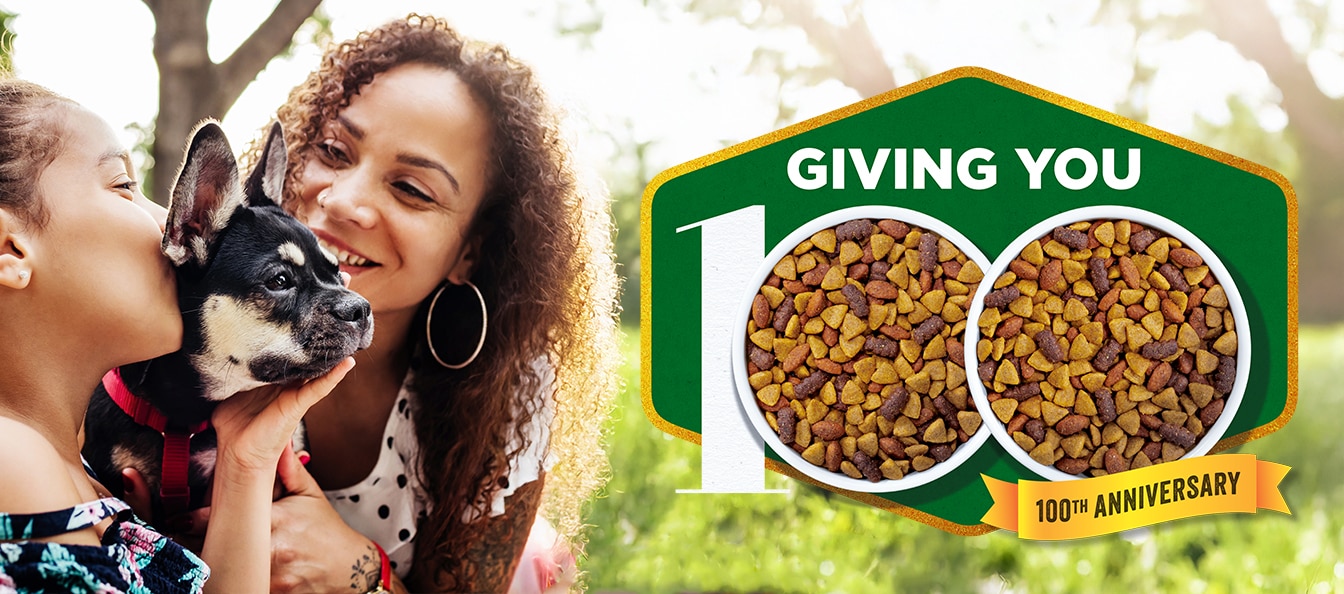 A woman and young girl show affection to a smal black and tan dog; the Dog Chow Giving You 100 logo sits to the right