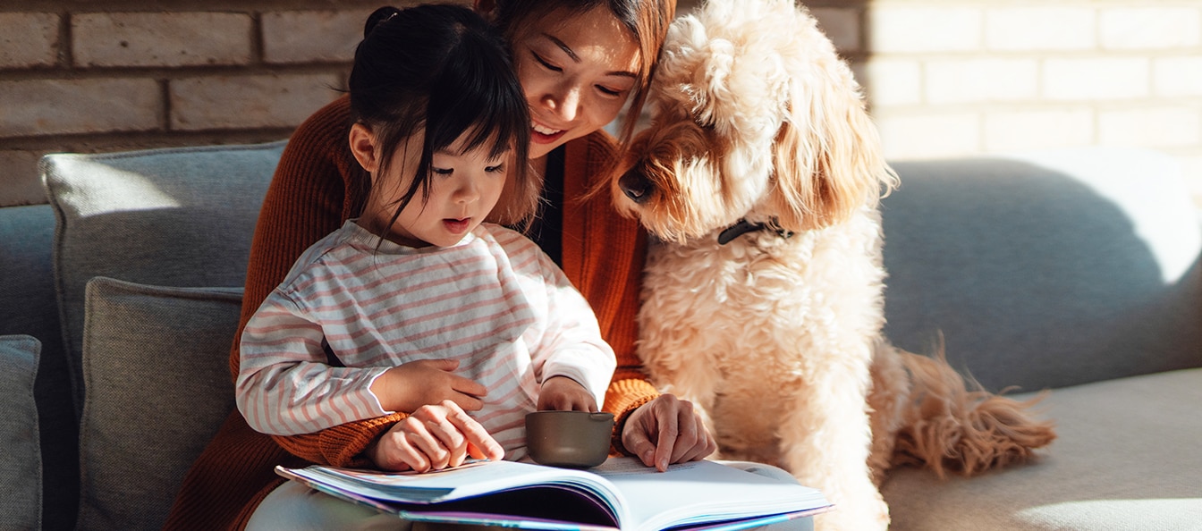 A woman reads to a small child sitting on her lap, while a curly-haired dog sits next to them on a couch